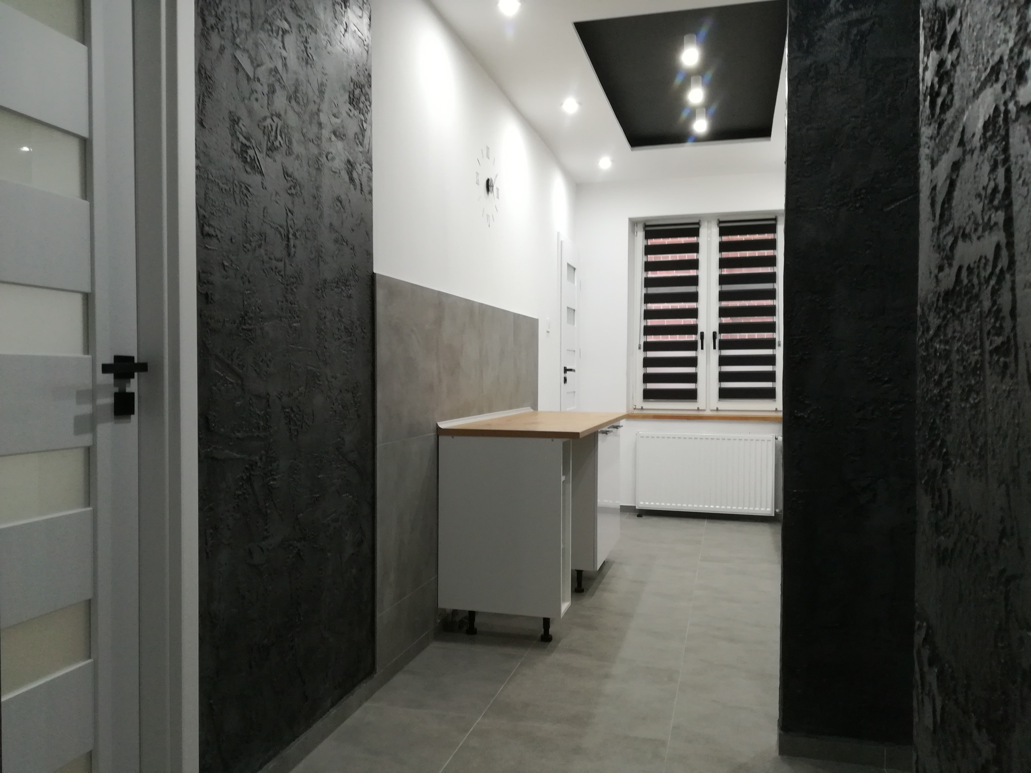 Nowoczesna kuchnia w trakcie montażu: białe szafki z drewnianym blatem, szare płytki podłogowe, ściany z dekoracyjnym tynkiem w odcieniach szarości i czerni, okno z roletami dzień-noc.