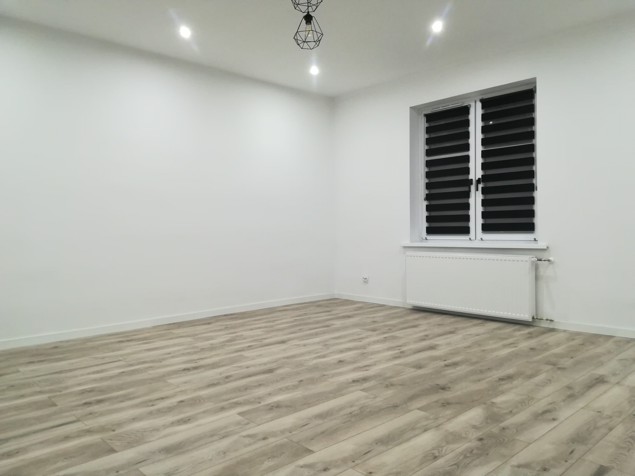Pusty, nowocześnie wykończony pokój z panelami podłogowymi imitującymi drewno, białymi ścianami, grzejnikiem pod oknem z żaluzjami i designerską lampą sufitową.