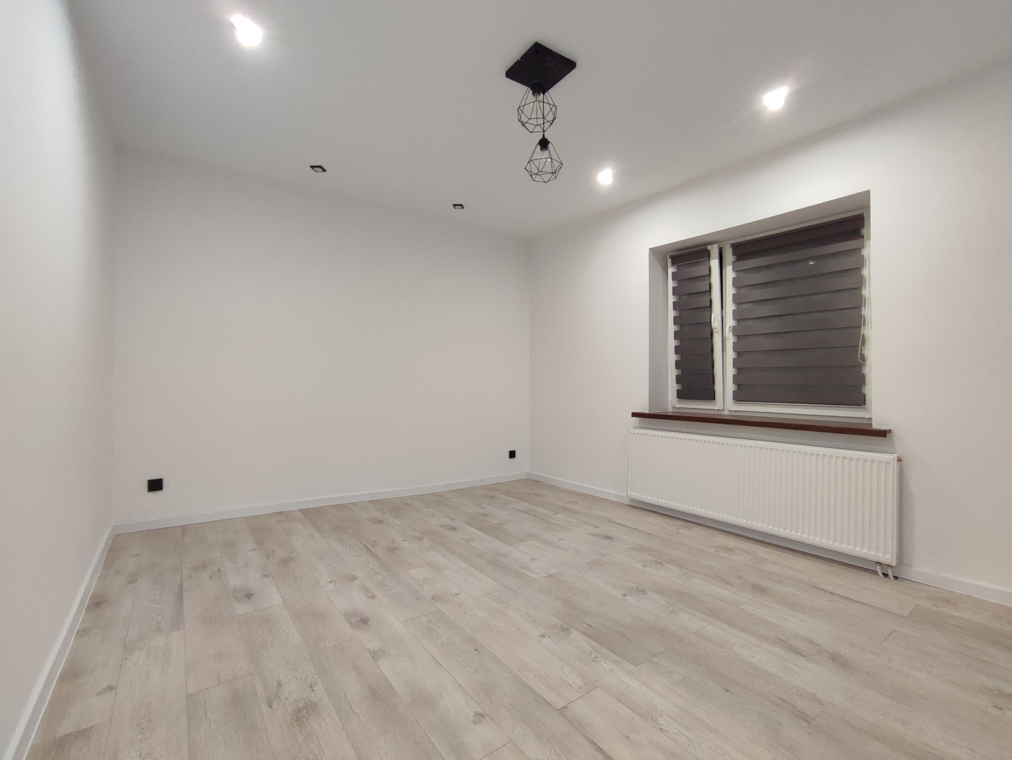 Pusty, odnowiony pokój z jasnymi panelami podłogowymi, białymi ścianami, grzejnikiem pod oknem z żaluzjami oraz nowoczesną lampą sufitową w stylu industrialnym.