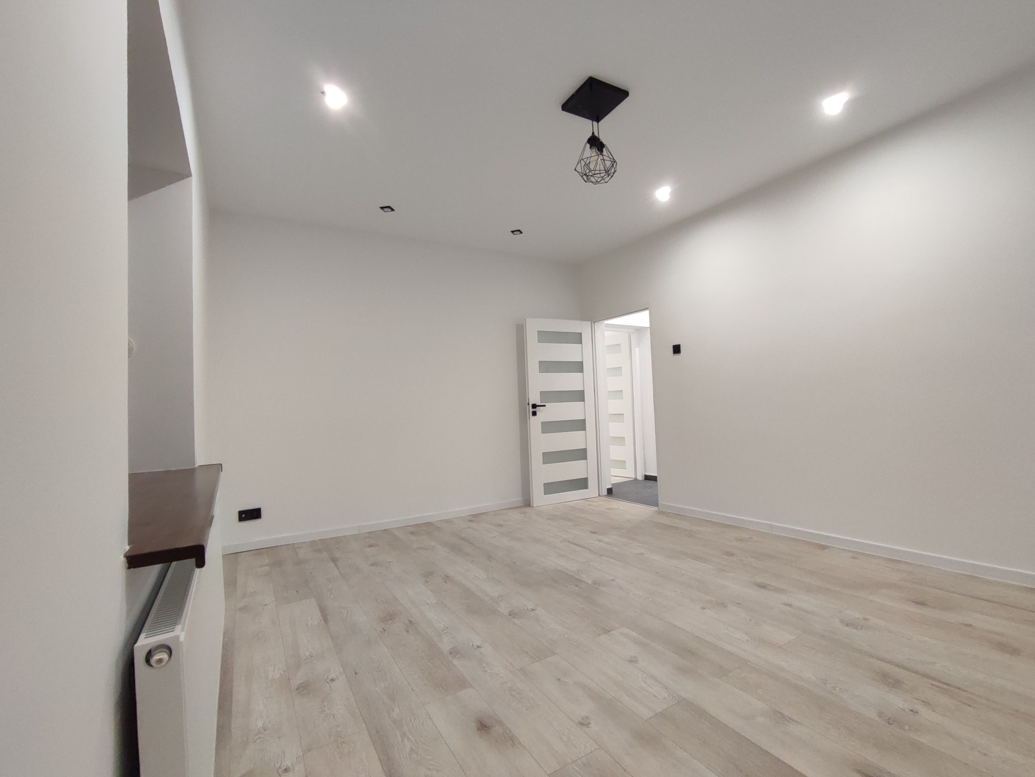 Pusty, nowo wykończony pokój z drewnopodobną podłogą, białymi ścianami, ozdobną lampą sufitową i drzwiami z przeszkleniami, gotowy do umeblowania.