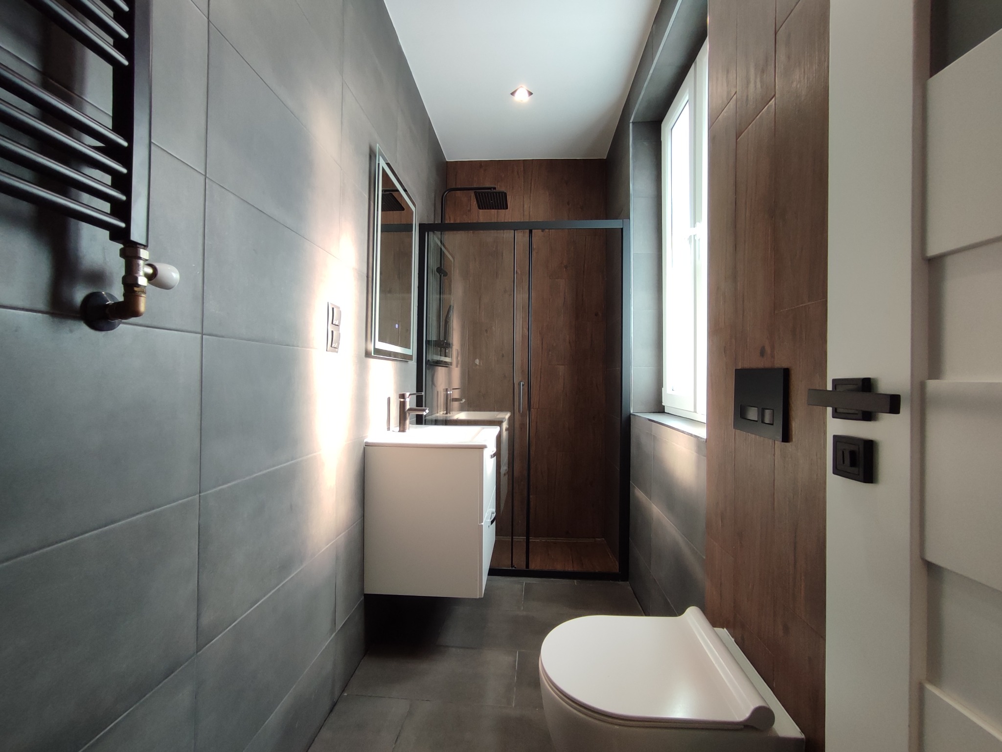 Nowoczesna łazienka z szarymi płytkami, kabiną prysznicową z czarnymi ramami, białym wiszącym blatem z umywalką i grzejnikiem drabinkowym.