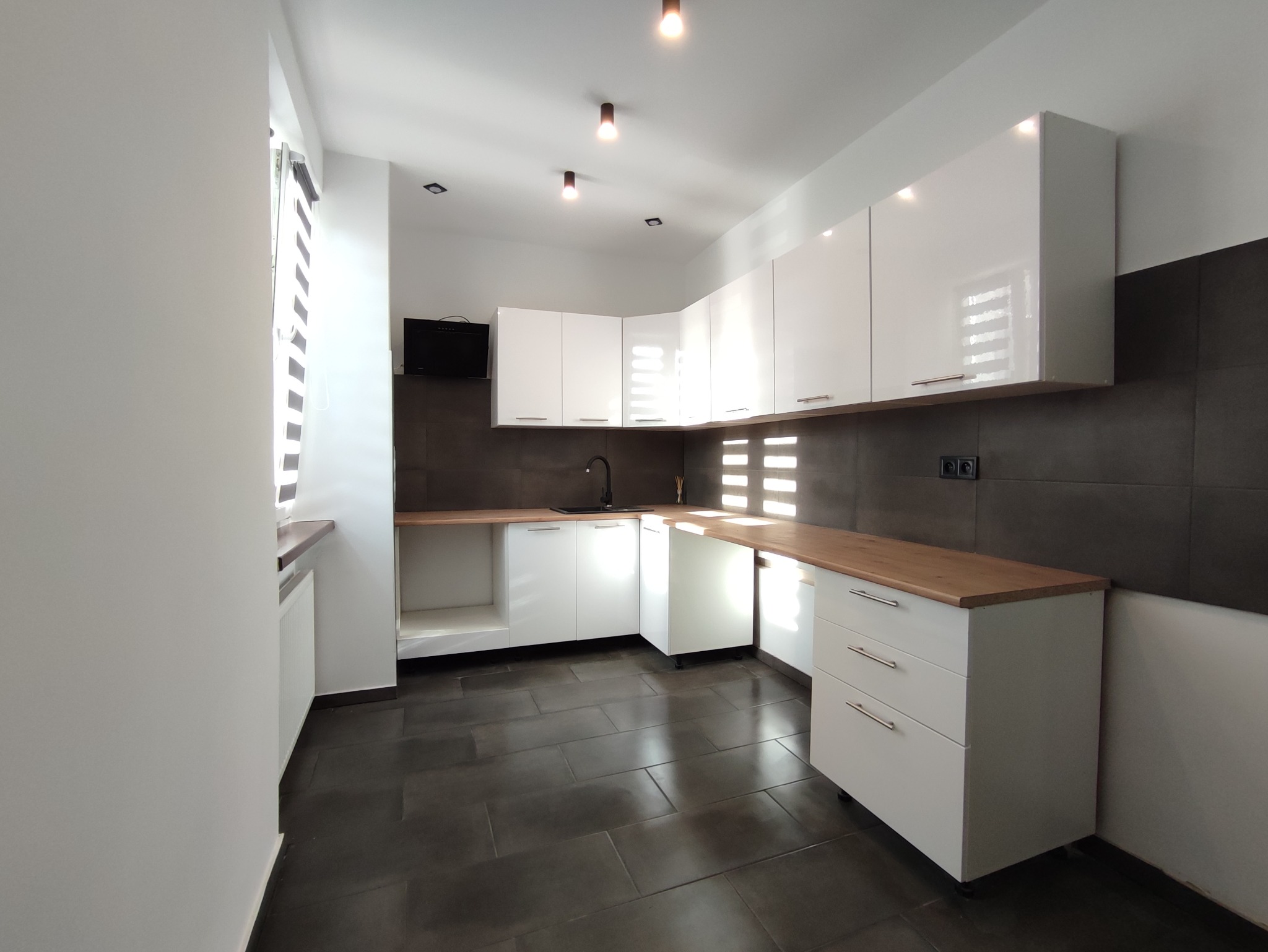 Nowoczesna kuchnia z białymi, lakierowanymi szafkami, drewnianym blatem i ciemnoszarą ścianą nad blatem, oświetlona trzema punktowymi lampami sufitowymi.