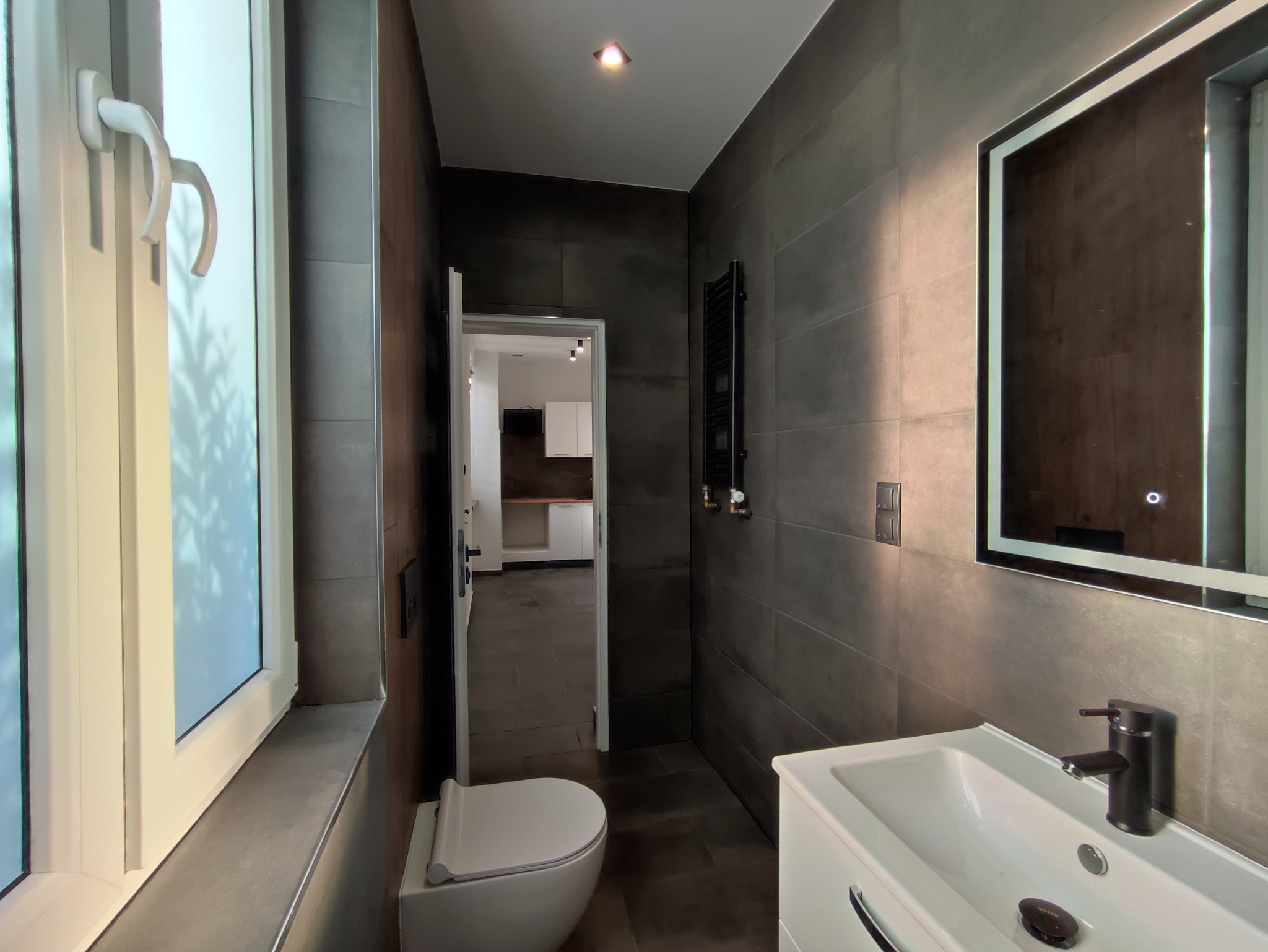 Nowoczesna łazienka z szarymi płytkami, podświetlanym lustrem i widokiem na kuchnię przez otwarte drzwi, z widocznym grzejnikiem drabinkowym i białą ceramiką.