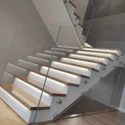 Nowoczesne schody z drewnianymi stopniami podświetlonymi LED, zabezpieczone szklaną balustradą mocowaną punktowo do stopni.