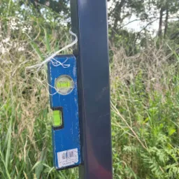 Ustawianie pionu słupka konstrukcyjnego za pomocą poziomicy, przymocowanej sznurkiem, widoczna ręka w czerwonej rękawicy roboczej.