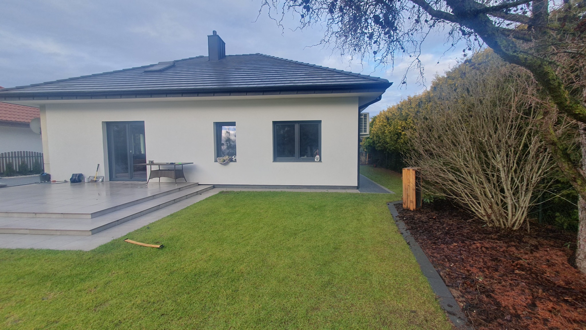 Nowoczesny dom jednorodzinny z białymi ścianami, ciemnoszarą dachówką i tarasem wyłożonym płytami na schodach, widok z trawnika w pochmurny dzień.