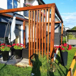 Nowoczesna drewniana pergola z pionowymi listwami, obok domu z balkonem, ozdobiona czerwonymi kwiatami w donicach, z widocznym cieniem osoby fotografującej.
