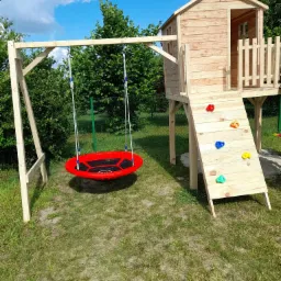 Drewniany plac zabaw dla dzieci z domkiem na palach, ścianką wspinaczkową z kolorowymi uchwytami i huśtawką typu bocianie gniazdo w kolorze czerwonym, ustawiony na trawniku w ogrodzie.