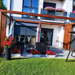 Drewniana konstrukcja z lamelami tworząca zadaszenie tarasu z meblami ogrodowymi i kwiatami w donicach, widok na nowoczesny dom z balkonem i roletami zewnętrznymi.