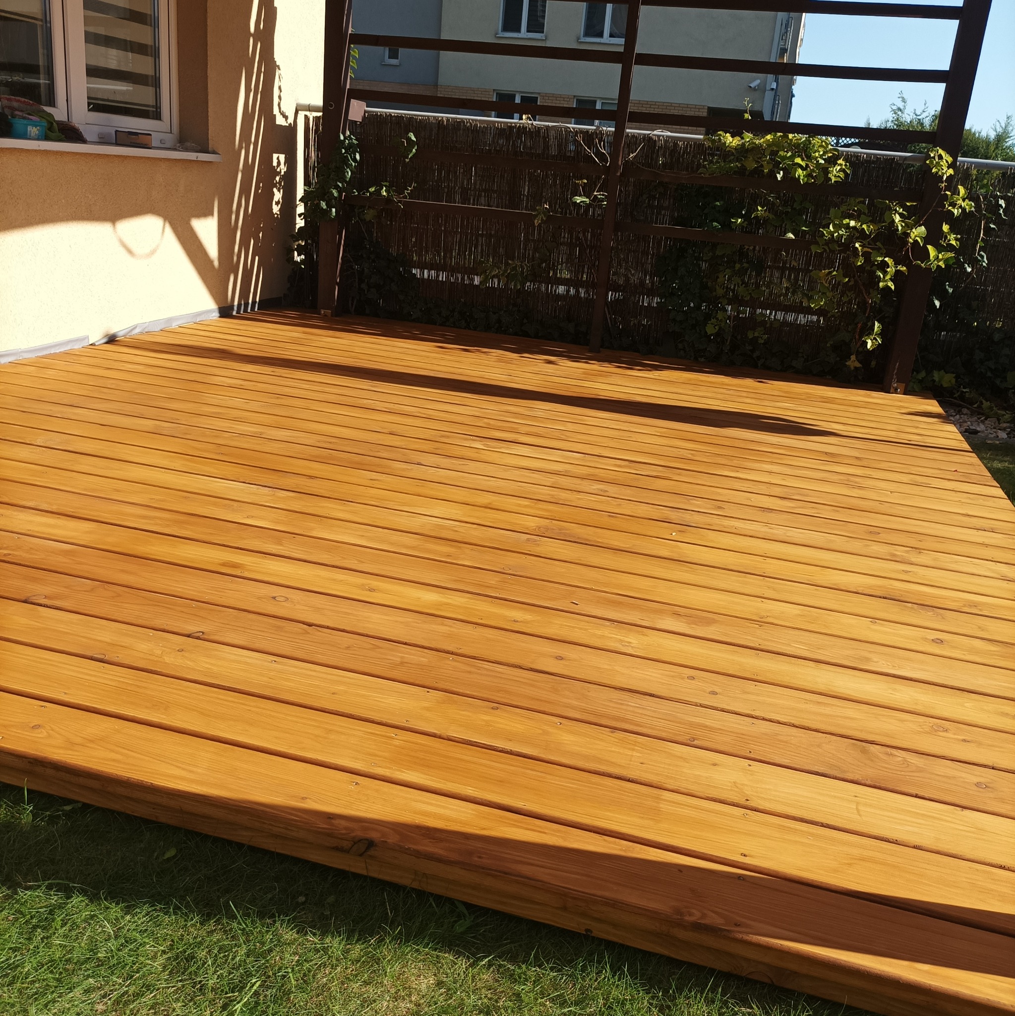 Nowy taras z desek kompozytowych w odcieniach miodowego brązu, z metalową pergolą częściowo porośniętą zielenią, przylegający do budynku.