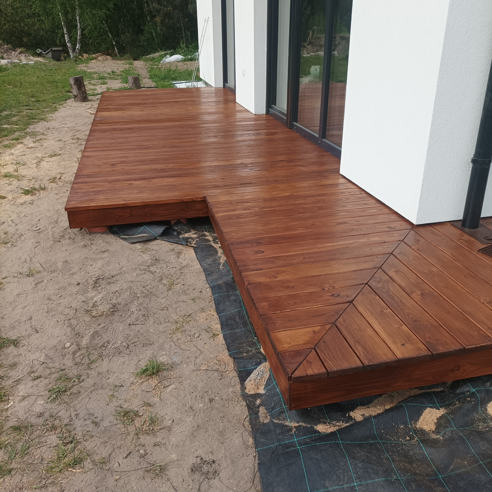 Świeżo polakierowany drewniany taras przylegający do białego domu z dużymi oknami, widoczna geowłóknina pod konstrukcją.