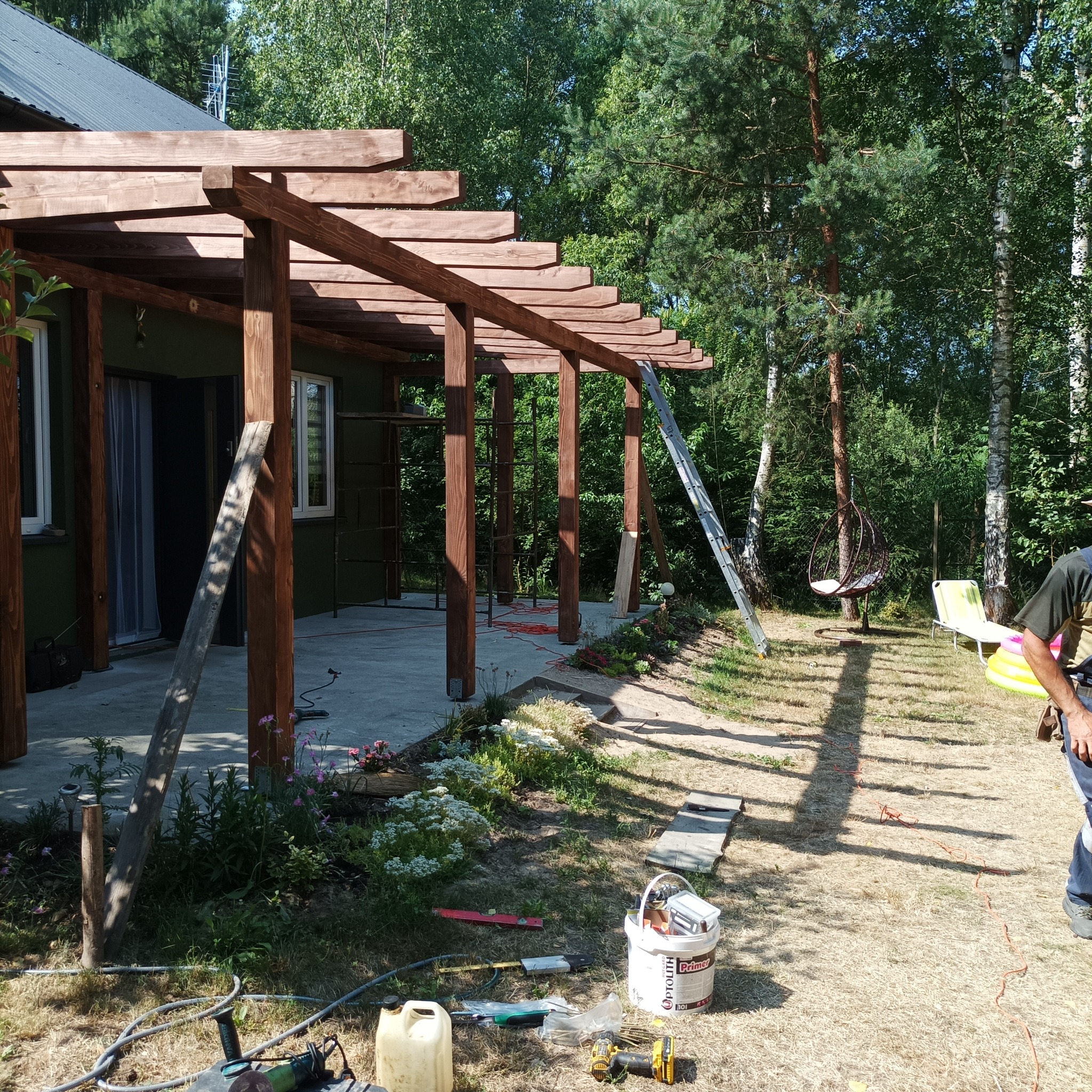 Drewniana pergola w trakcie budowy przy zielonym domu, widoczne narzędzia i materiały budowlane, drabina oparta o konstrukcję, w tle drzewa.