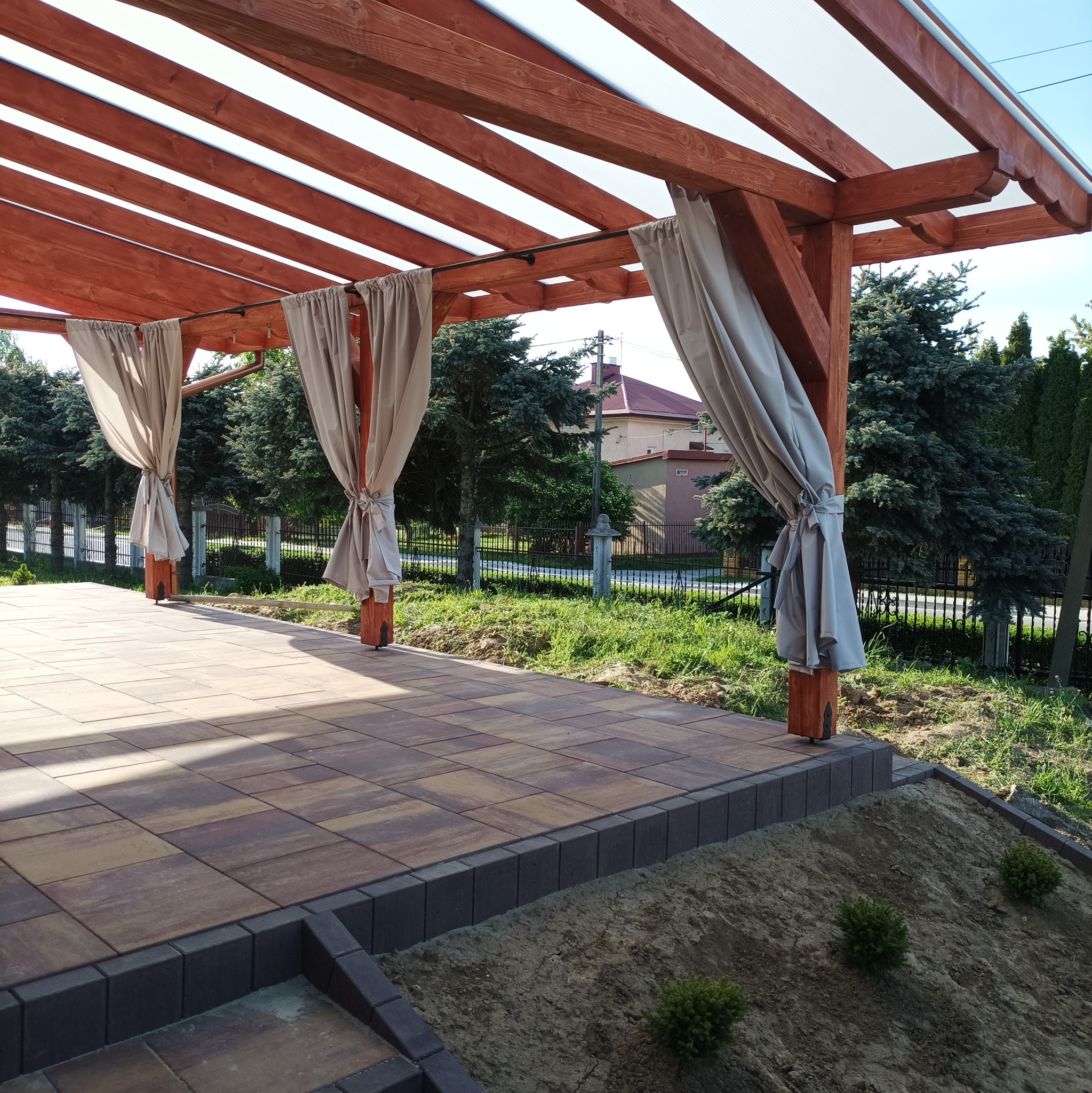 Drewniana pergola z jasnymi zasłonami na metalowym karniszu, zadaszona przezroczystym tworzywem, z brukowanym tarasem i widokiem na ogród z tujami i domem w tle.