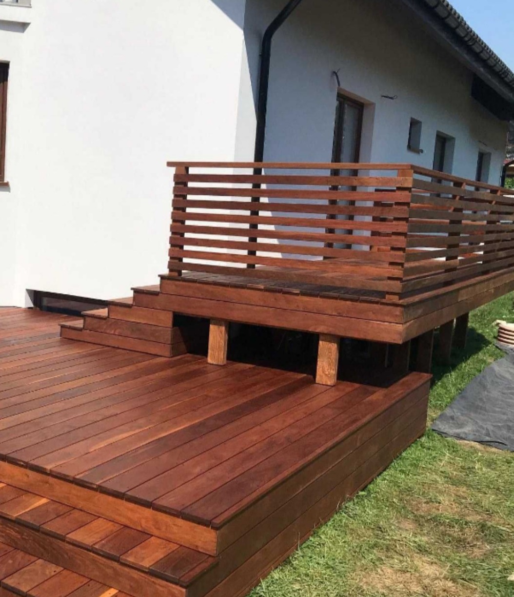 Nowoczesny taras drewniany z poziomymi deskami balustrady, schodami i widocznymi podporami, przylegający do białego budynku z oknami.