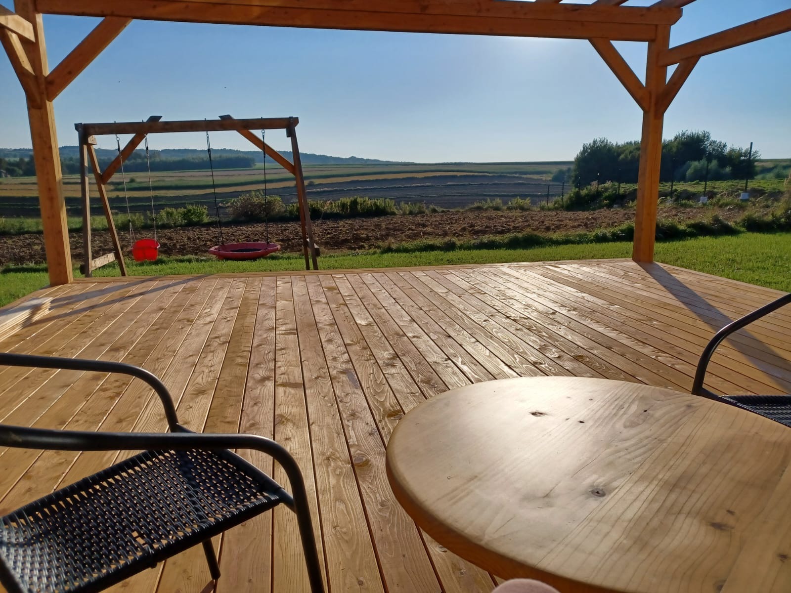 Drewniany taras z widokiem na pola, z drewnianą pergolą, okrągłym stołem i czarnym krzesłem z plecionki. W tle huśtawka ogrodowa z czerwonym siedziskiem.