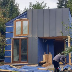 Gretter sp z oo firma elektryczno budowlana - Wznoszenie nowoczesnego domu modułowego z szarą elewacją i drewnianymi oknami, widoczne warstwy izolacyjne oraz pracownik w trakcie montażu, otoczenie z drzewami.