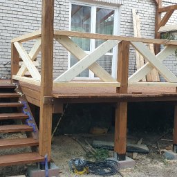 Gretter sp z oo firma elektryczno budowlana - Drewniany taras w trakcie budowy: widoczne brązowe deski, schody zabezpieczone niebieskimi taśmami, surowe drewniane balustrady, drabina oparta o ceglany budynek w tle.