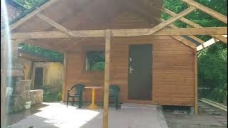 Niewielka, drewniana konstrukcja z płaskim dachem w trakcie budowy, widoczne elementy szkieletu dachu, z boku mały kamienny grill, obok stolik i krzesła.