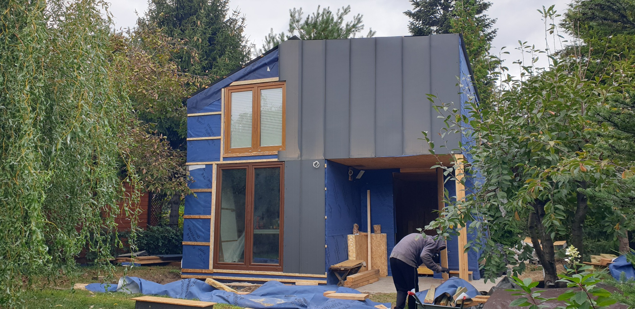 Wznoszenie nowoczesnego domu modułowego z szarą elewacją i drewnianymi oknami, widoczne warstwy izolacyjne oraz pracownik w trakcie montażu, otoczenie z drzewami.