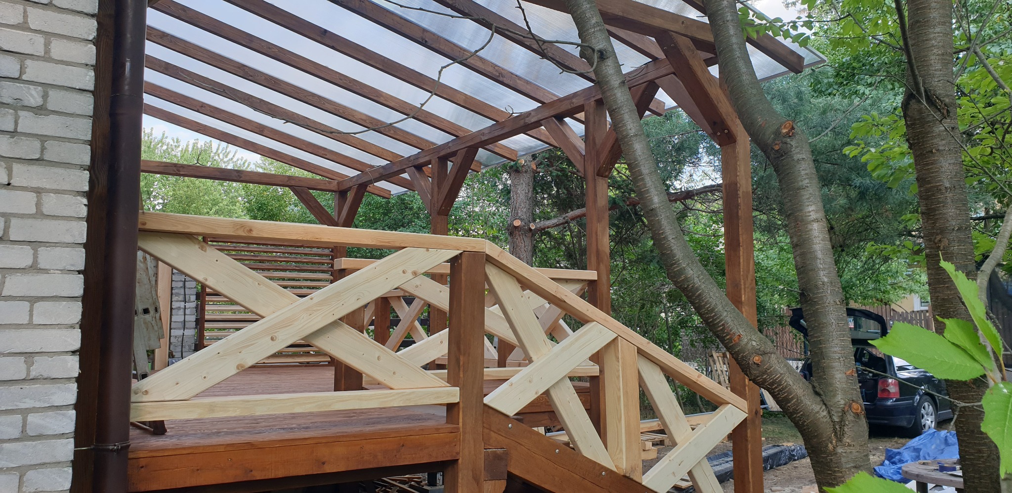 Zadaszona konstrukcja z drewna z balustradą w kształcie krzyża, widoczna z bliska, z drzewami w tle i fragmentem ściany budynku z boku.