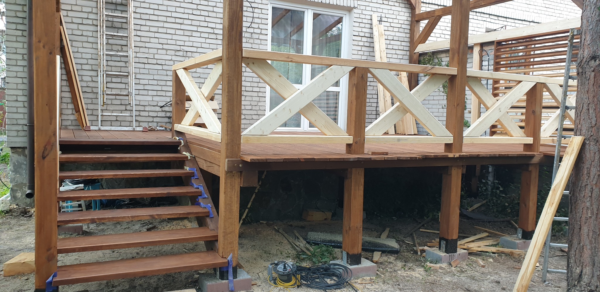 Drewniany taras w trakcie budowy: widoczne brązowe deski, schody zabezpieczone niebieskimi taśmami, surowe drewniane balustrady, drabina oparta o ceglany budynek w tle.