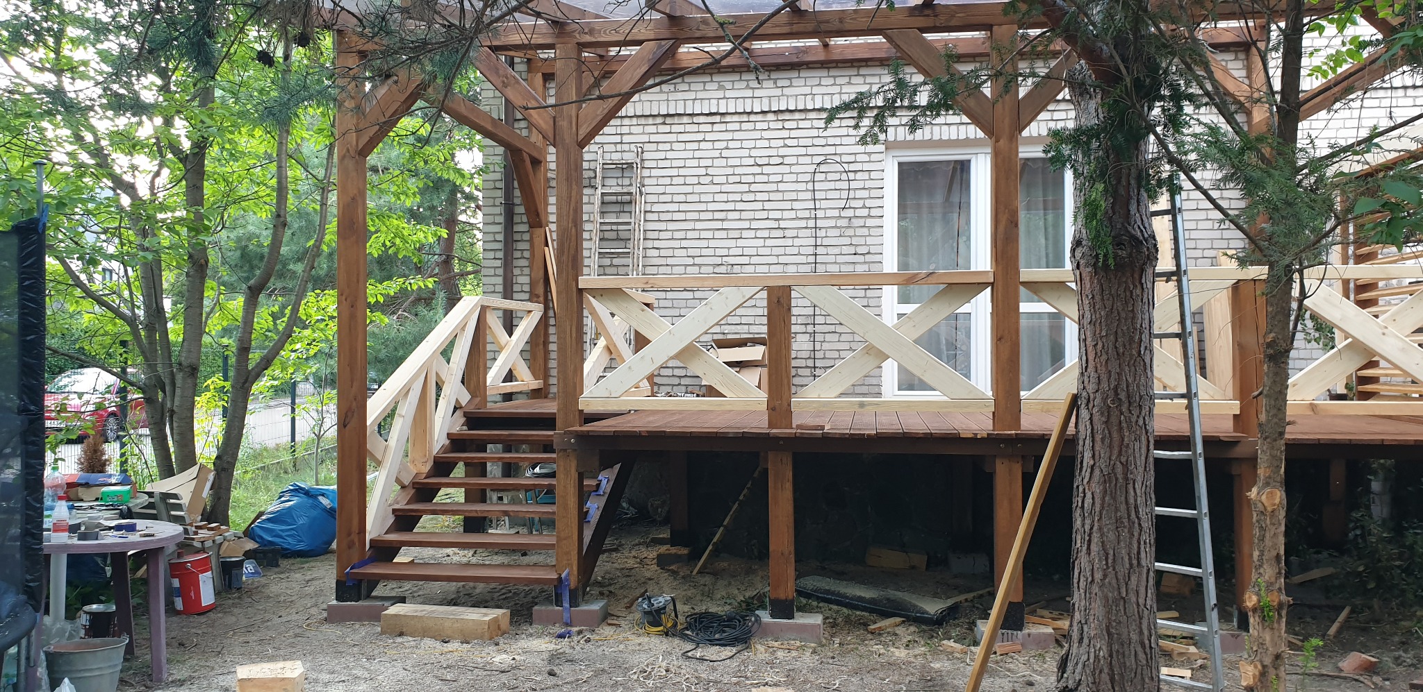 Drewniany taras z pergolą w trakcie budowy, widoczne schody, balustrady z krzyżowym wzorem, narzędzia i materiały budowlane wokół.