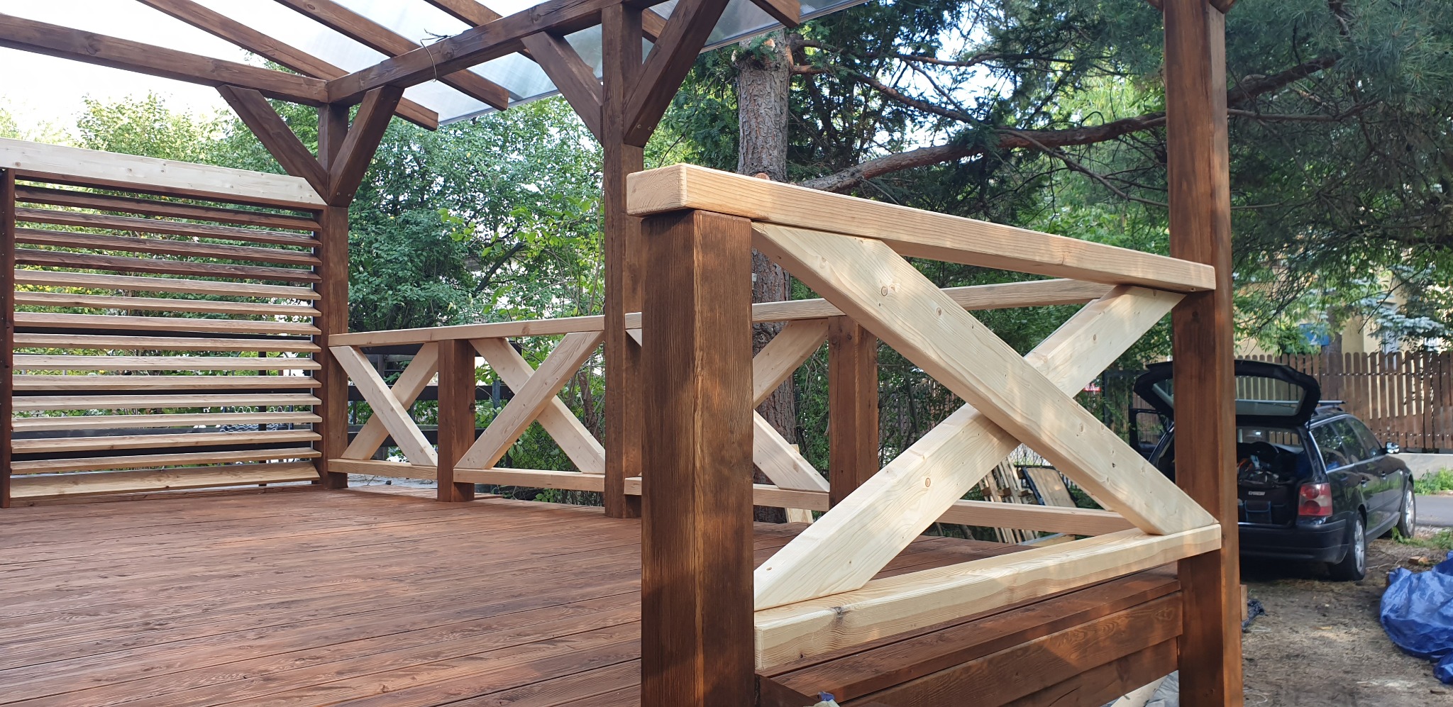 Zadaszona konstrukcja drewniana z podłogą z desek, częściowo pomalowana na brązowo, z elementami kratownicy i żaluzji, widok na zieleń i zaparkowany samochód.