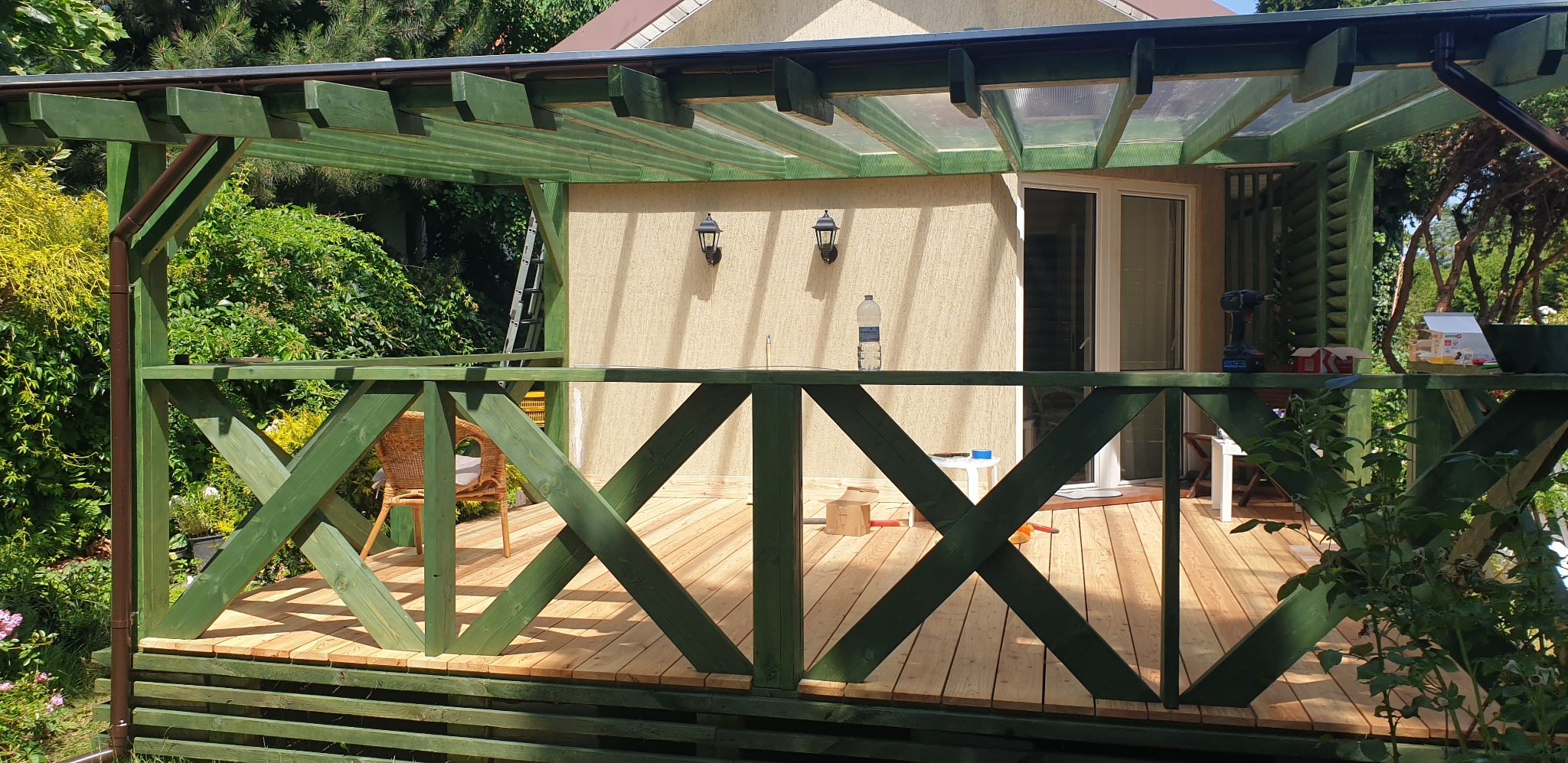 Zadaszony taras z drewnianą balustradą pomalowaną na zielono, widoczne narzędzia i materiały budowlane, krzesło wiklinowe i roślinność w tle. Częściowo ukończony projekt.