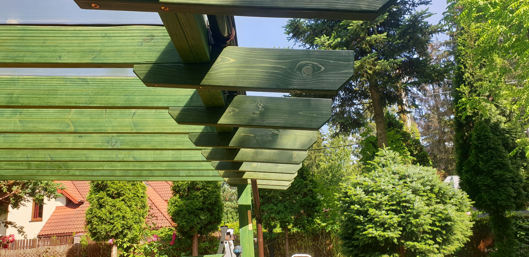 Widok spod spodu zielonej, drewnianej konstrukcji z elementami przezroczystego zadaszenia, w tle zieleń drzew i fragment dachu domu.