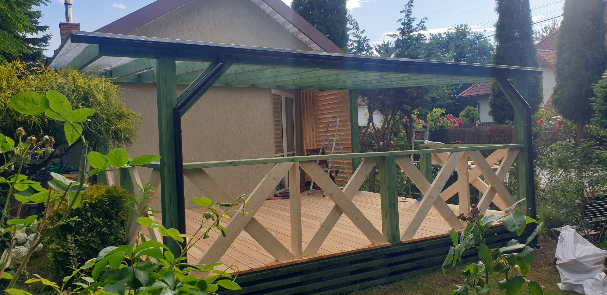 Drewniana pergola z zielonymi elementami konstrukcyjnymi i jasną drewnianą podłogą tarasu w trakcie budowy, widoczne narzędzia, dom w tle, roślinność ogrodowa.