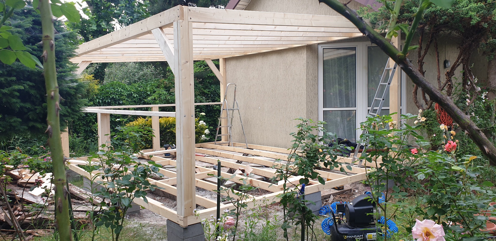 Budowa drewnianej konstrukcji przy budynku, widoczne elementy nośne i podłogowe, drabina oparta o ścianę, kompresor pneumatyczny na pierwszym planie, otoczenie ogrodowe z różami.