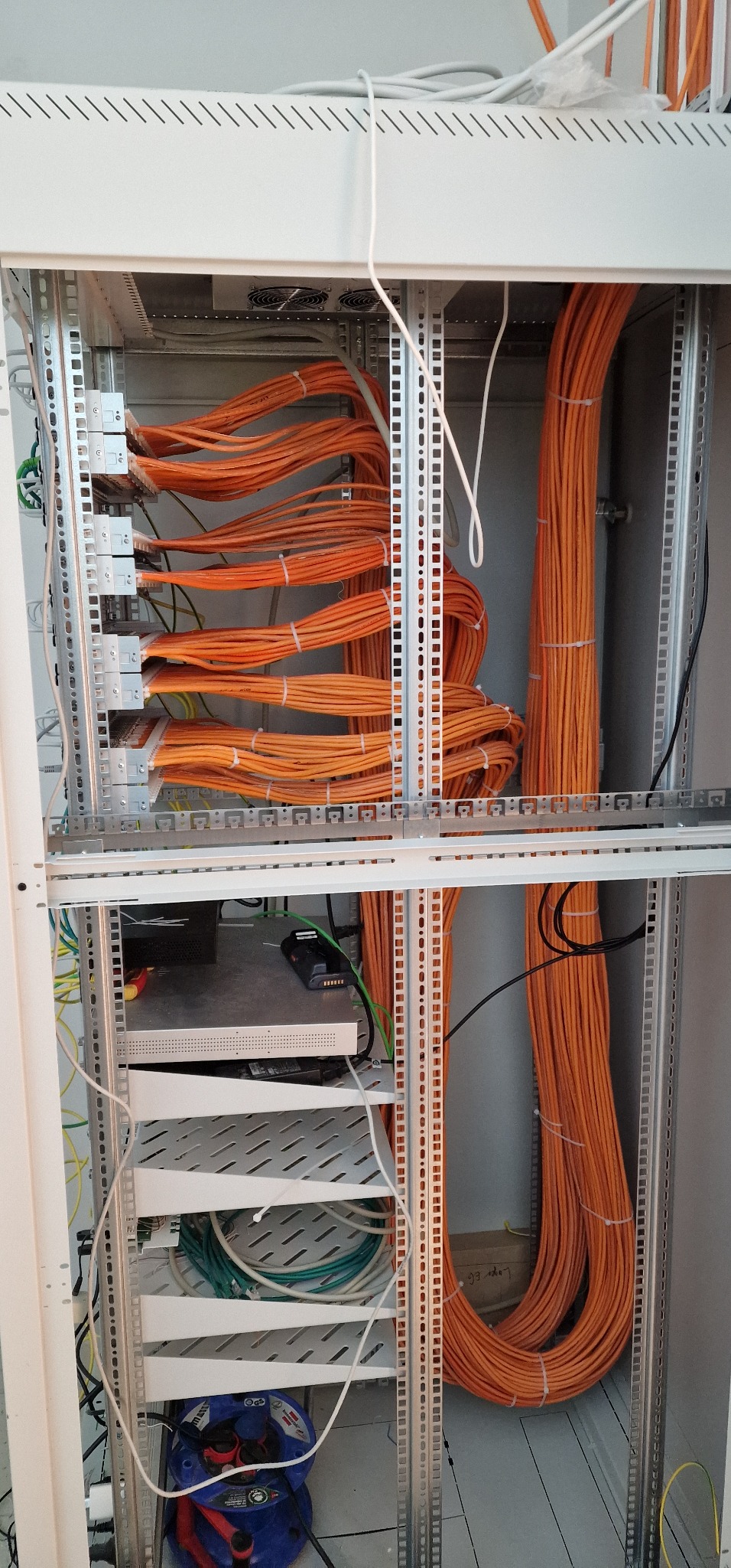 Szafa serwerowa z uporządkowanymi pomarańczowymi kablami sieciowymi, widoczne panele krosownicze i zwinięty przedłużacz na dole szafy.