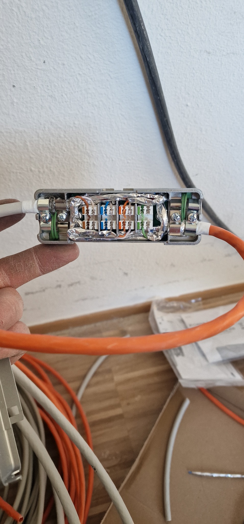 Otwarta metalowa puszka połączeniowa z widocznymi złączkami kablowymi, zabezpieczona folią aluminiową, trzymana w palcach, z widocznymi kablami w kolorach pomarańczowym i białym w tle.