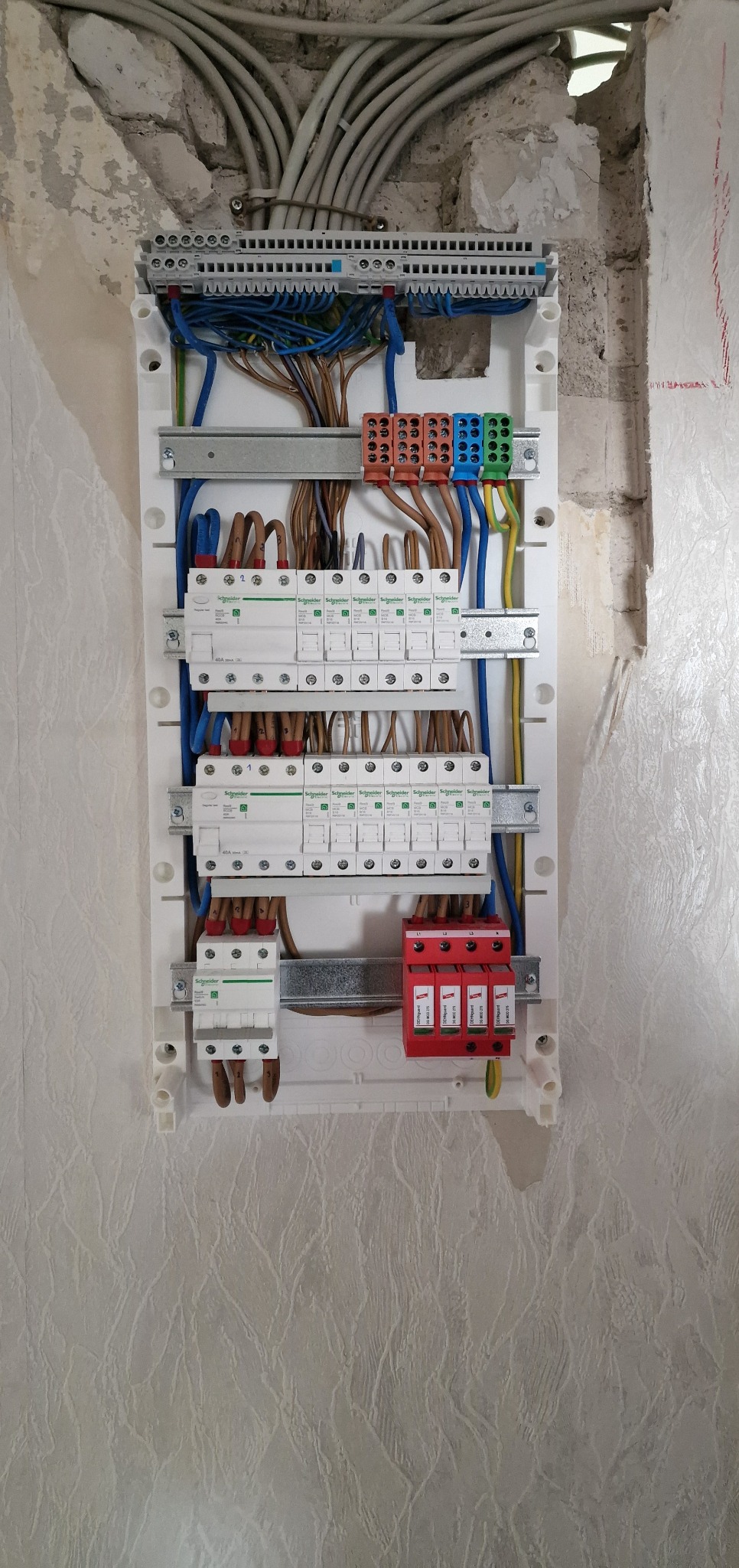Rozdzielnica elektryczna Schneider z zamontowanymi zabezpieczeniami, widoczne kable w kolorowych izolacjach i listwy zaciskowe, montaż na ścianie z nierówną fakturą.