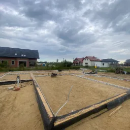 Przygotowanie fundamentów domu jednorodzinnego: wykop z obrysem, szalunki z folią kubełkową, poziomica laserowa, w tle budynek w stanie surowym otwartym pod pochmurnym niebem.