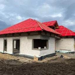 Wznoszenie ścian domu z betonu komórkowego w Namysłowie, widoczna konstrukcja dachu z czerwoną membraną i drewnianymi łatami.
