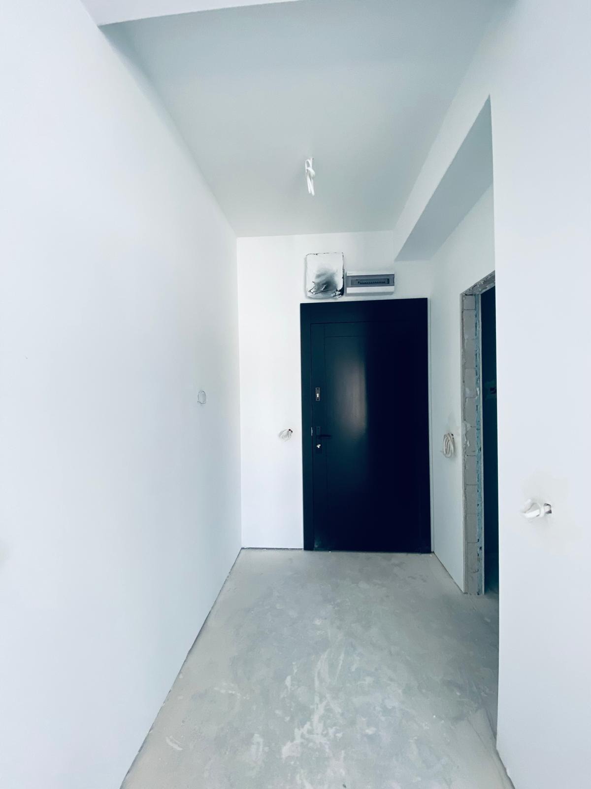 Białe wnętrze korytarza z czarnymi drzwiami wejściowymi i surowym betonem na podłodze, widoczne przygotowane punkty elektryczne na ścianach.