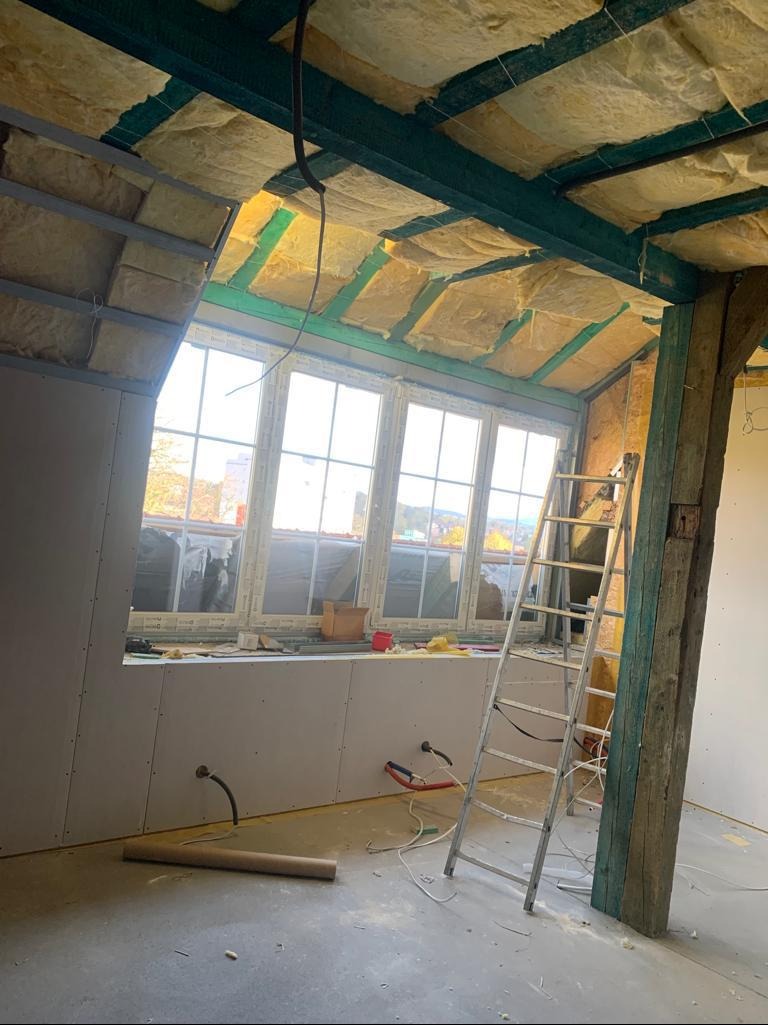 Poddasze w trakcie wykańczania: izolacja wełną mineralną, częściowo zamontowane płyty gipsowo-kartonowe, drabina oparta o drewniany słup. Widok na okno.