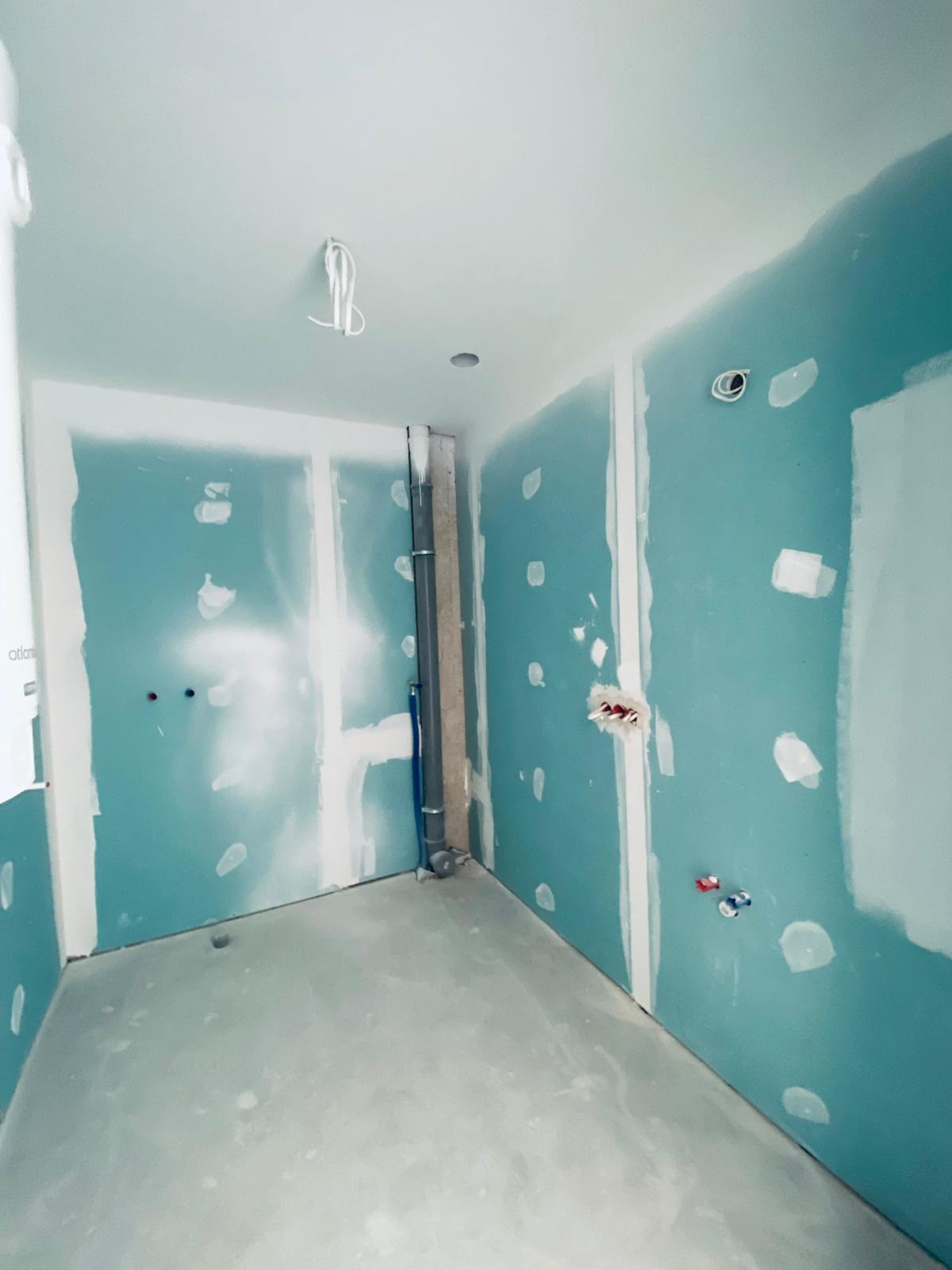 Wnętrze w trakcie wykańczania: ściany z płyt karton-gips, widoczne instalacje wodne i elektryczne, surowy beton na podłodze. Prace wykończeniowe w łazience.