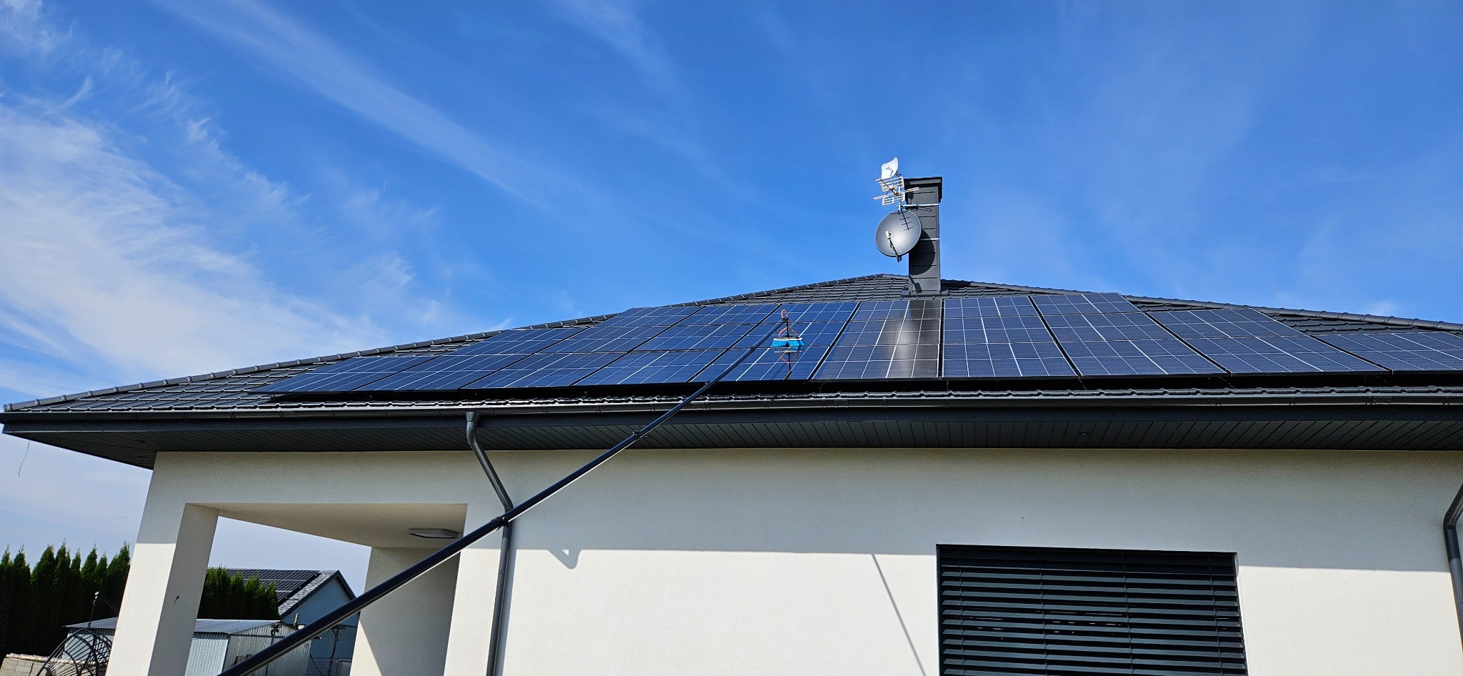 Czyszczenie paneli słonecznych na dachu domu jednorodzinnego za pomocą teleskopowej szczotki, widoczny komin z antenami satelitarnymi na tle błękitnego nieba z chmurami.