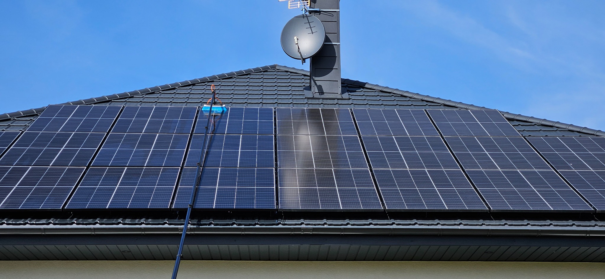 Czyszczenie paneli słonecznych na dachu domu za pomocą szczotki na długim teleskopowym kiju. Widoczna różnica w czystości między umytą a nieumytą częścią panelu.