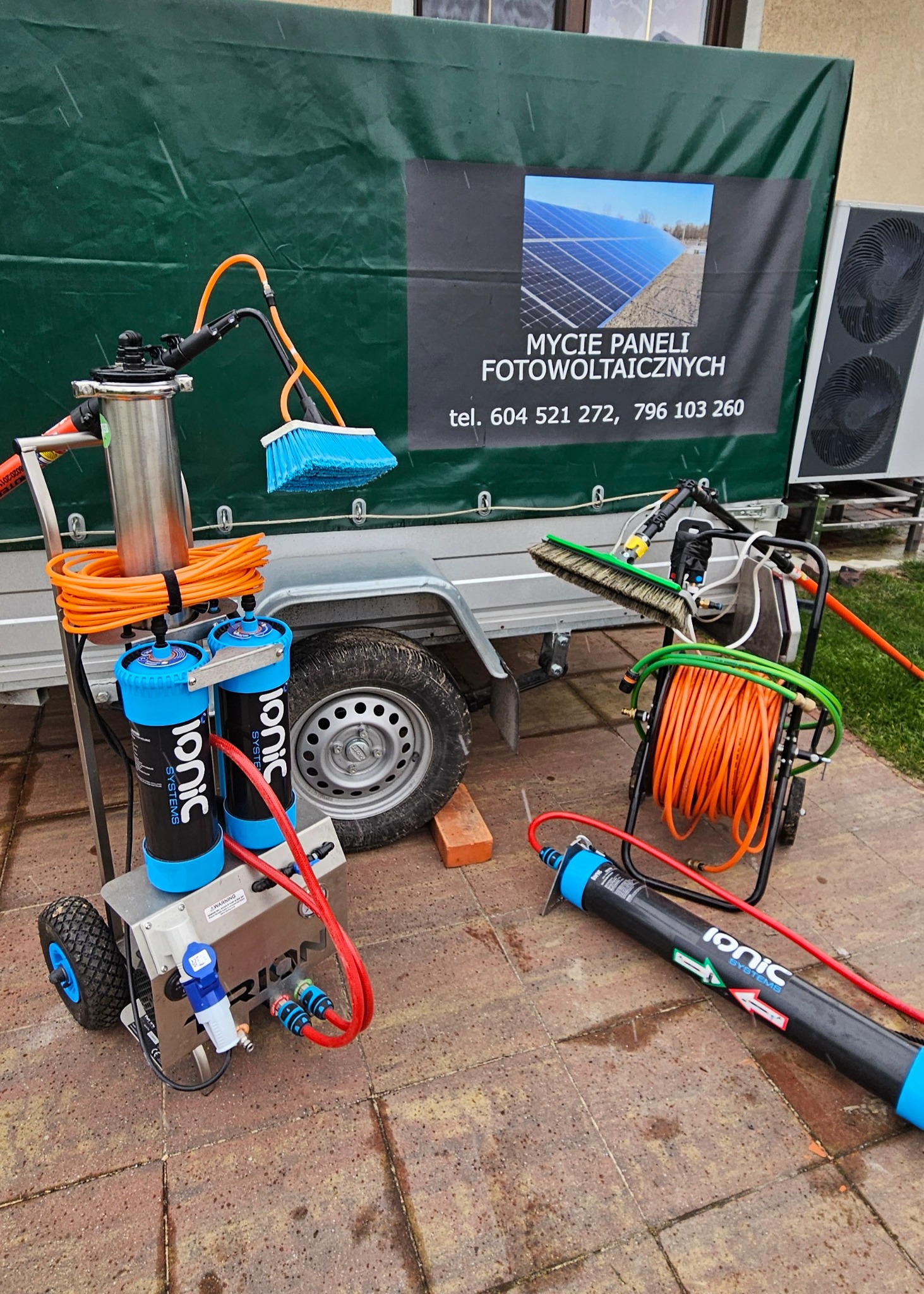 Mobilne urządzenia do czyszczenia paneli fotowoltaicznych, z widoczną szczotką, wężami i filtrami Ionic Systems, na tle przyczepki z banerem reklamowym 'Mycie paneli fotowoltaicznych' i numerami...