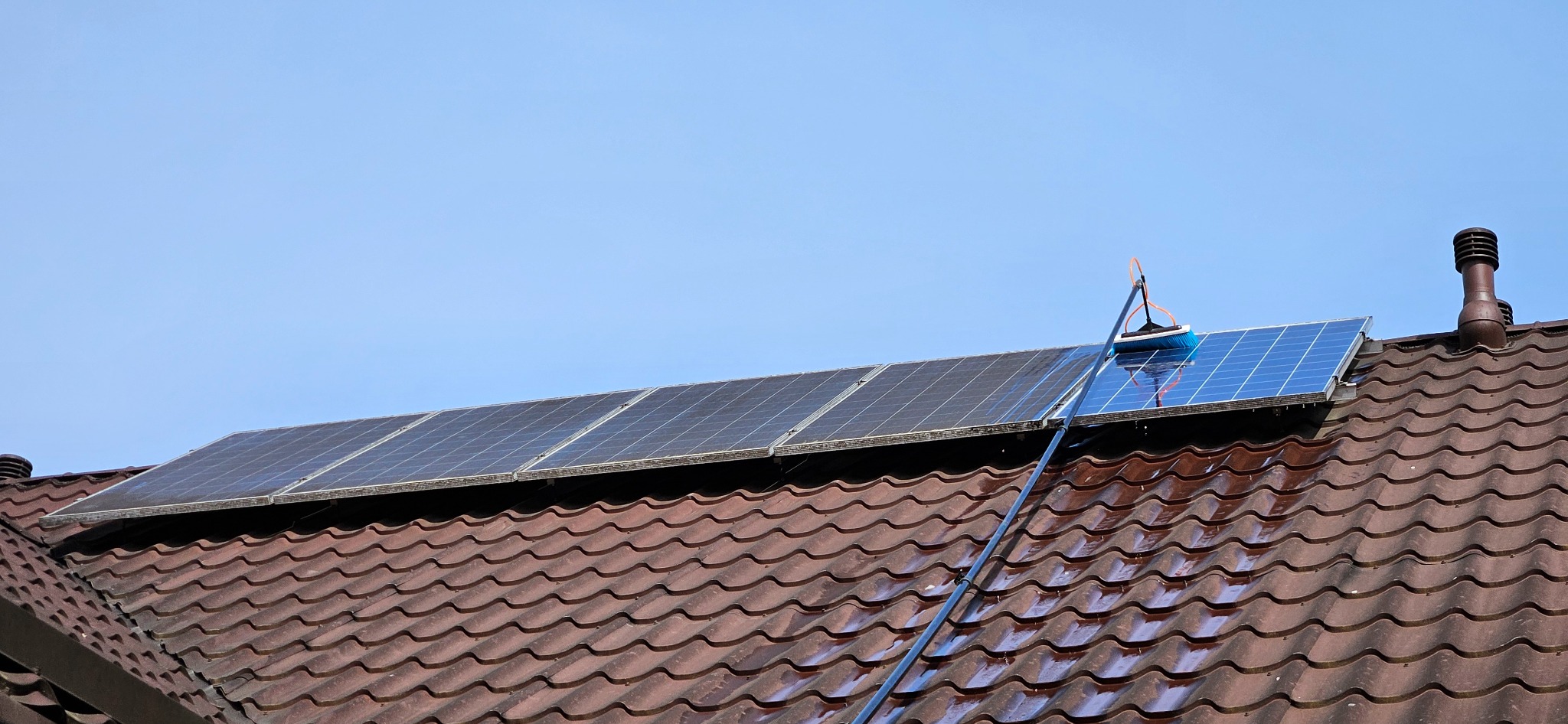 Czyszczenie paneli słonecznych na dachu z brązowej dachówki za pomocą szczotki na teleskopowym kiju, błękitne niebo w tle.