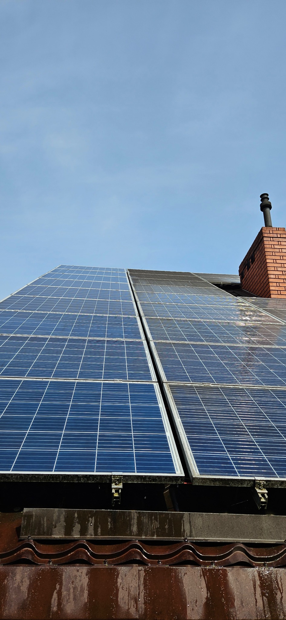 Panele słoneczne na dachu z brązowej dachówki, obok ceglany komin z czarnym kominkiem, widoczne ślady po deszczu.