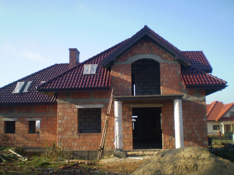 Niedokończony budynek jednorodzinny z czerwoną dachówką, widoczne pustaki i brak okien, z drabina opartą o ścianę.