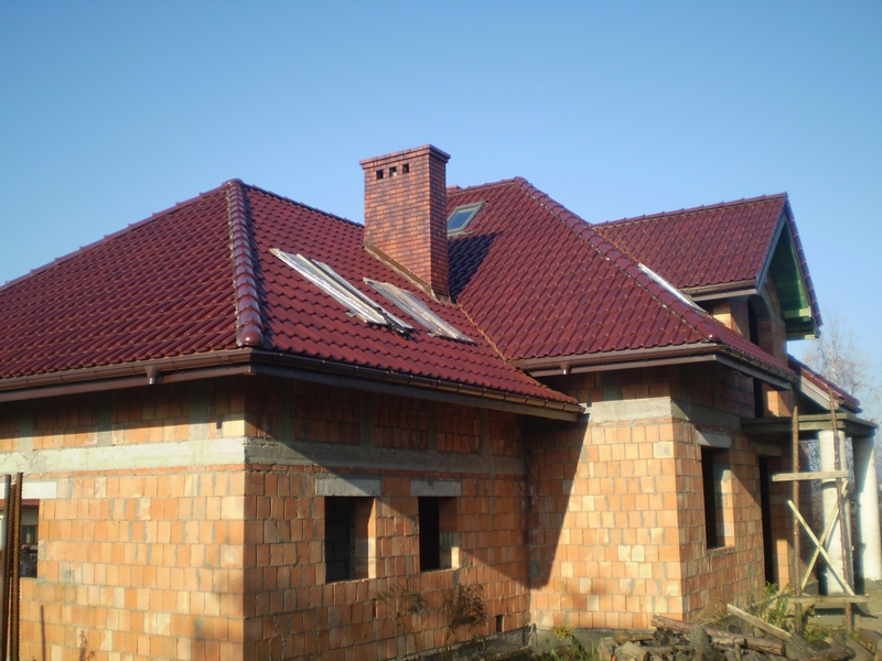 Dom w trakcie budowy z nowym, czerwonym dachem z dachówki ceramicznej, widoczne rynny i komin z cegły, częściowo zabezpieczony dach folią.