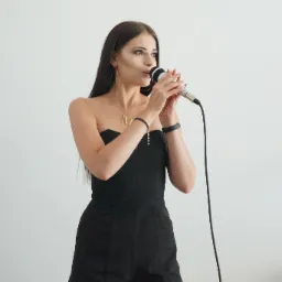 Kobieta w czarnym kombinezonie bez ramiączek, z długimi ciemnymi włosami, śpiewa do mikrofonu; widoczny złoty naszyjnik z literą 'M'.