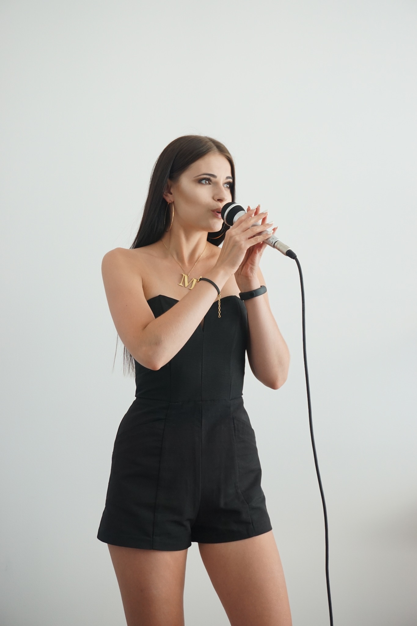 Kobieta w czarnym kombinezonie bez ramiączek, z długimi ciemnymi włosami, śpiewa do mikrofonu; widoczny złoty naszyjnik z literą 'M'.