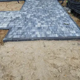 Świeżo ułożona szara kostka brukowa prowadząca do budynku, widoczny poziomica na kostce, z piaskowym podłożem w tle.