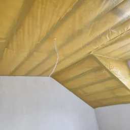 Poddasze w trakcie ocieplania, widoczna żółta wełna mineralna zabezpieczona folią, wystający kabel elektryczny, surowe ściany, okno w tle.